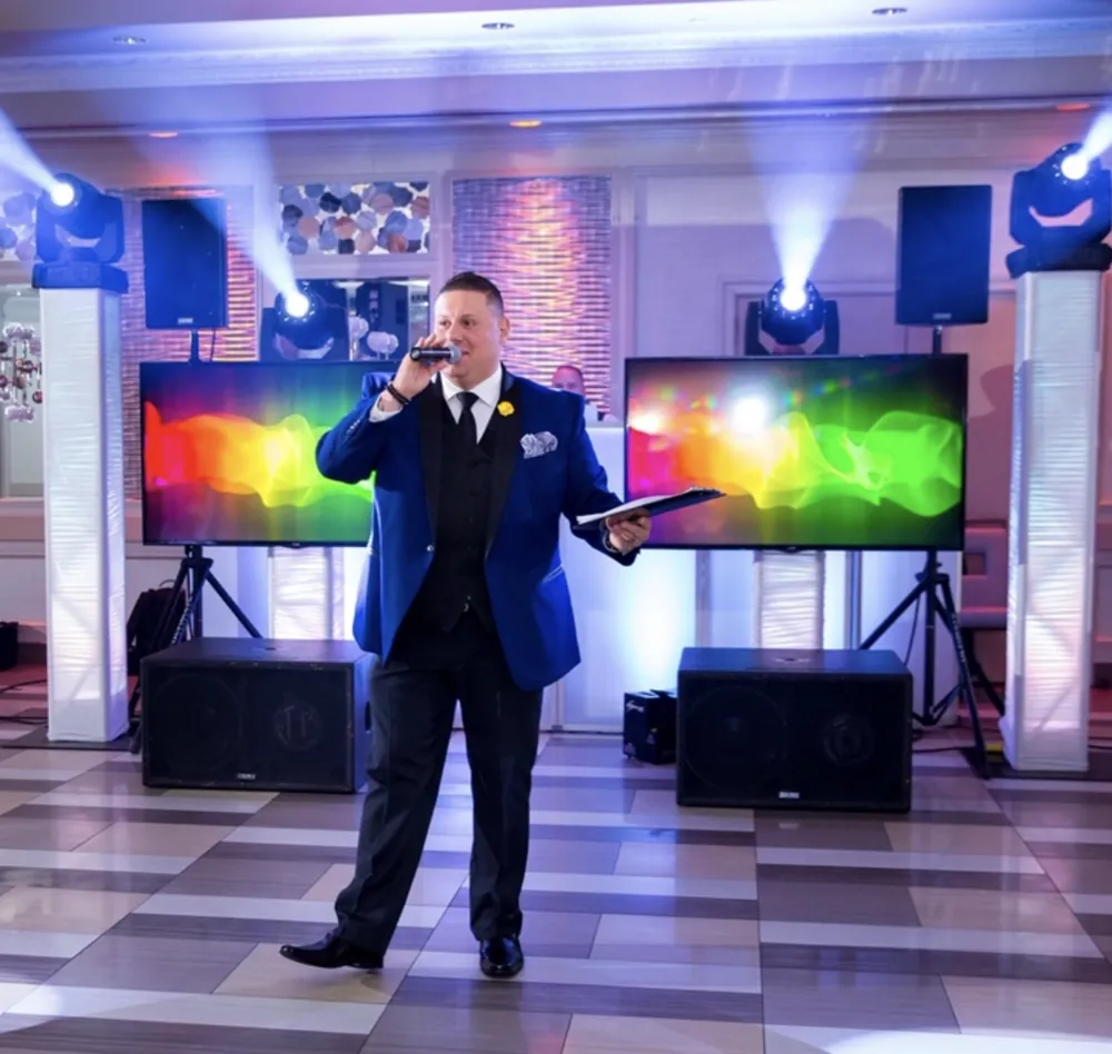 Wedding MC in a blue jacket speaking into a microphone in front of a DJ setup with video screens and moving lights
