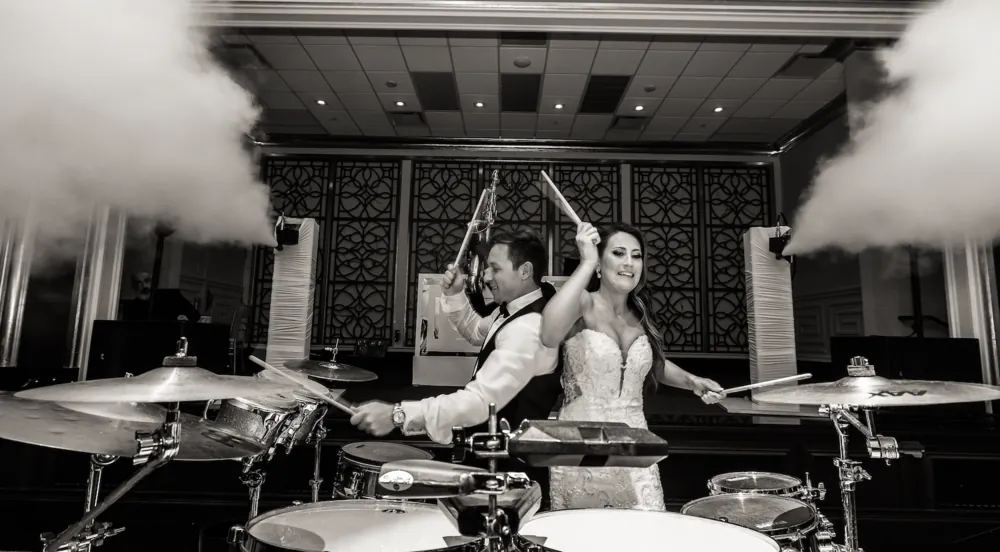 Black-and-white photo of a bride and groom playing drums as CO2 blast jets shoot up on each side