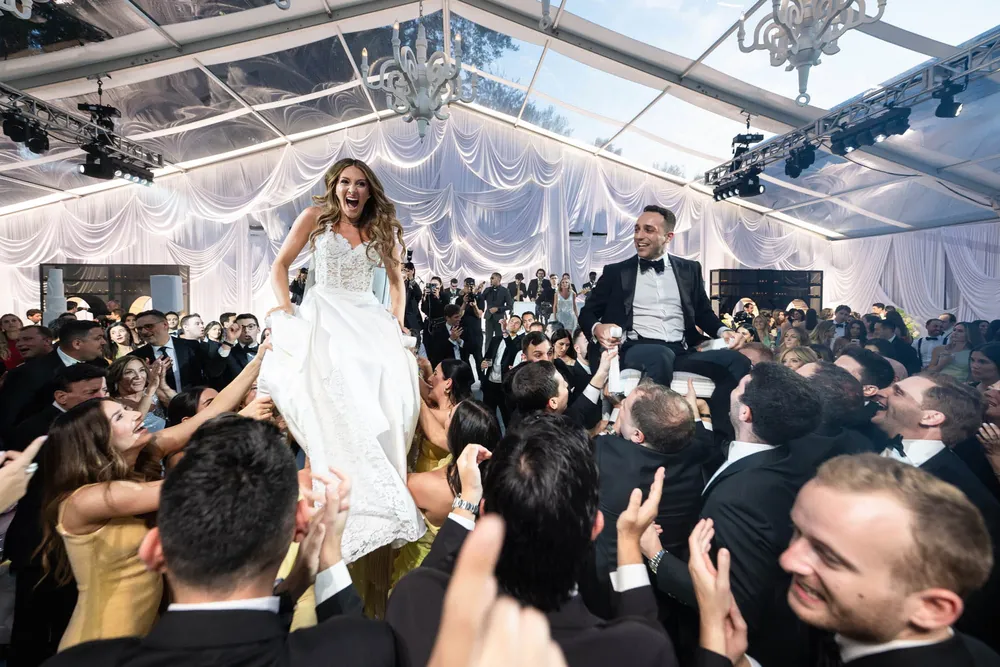 Bride and groom lifted on chairs by guests inside a clear wedding tent lit with stage lights and chandeliers