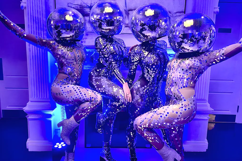 Four dancers in sequined bodysuits wearing mirrored disco ball helmets posing under blue light