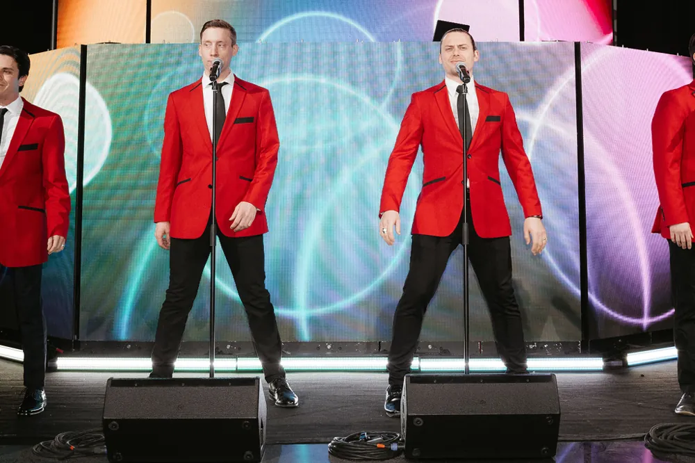 Singers in matching red jackets performing on stage in front of a colorful LED wall, a specialty act