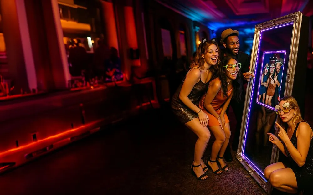 Guests in party glasses posing in front of a mirror photo booth with a light-up border and countdown