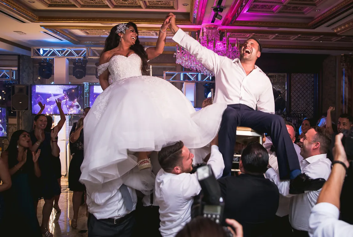 Bride and groom lifted on chairs for the hora as guests cheer at a lively wedding reception