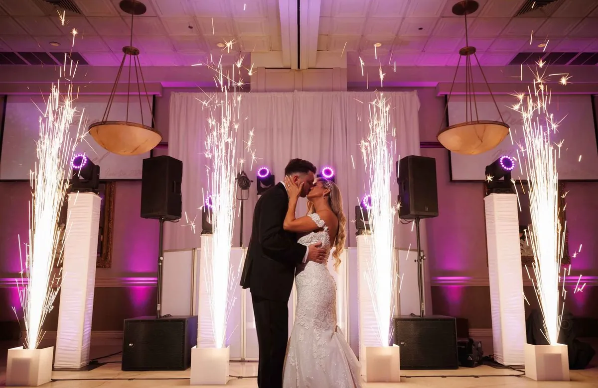 Bride and groom kissing between cold spark fountains and speakers under purple uplighting at a wedding reception