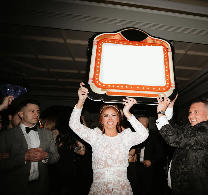 Bride holding a lit club marquee sign up over the dance floor as guests cheer around her