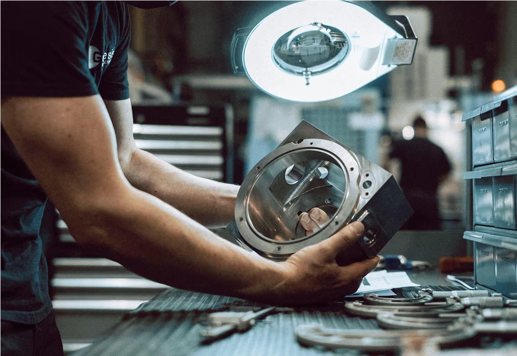 A man in a fabrication workshop holding a steel mechanical component.