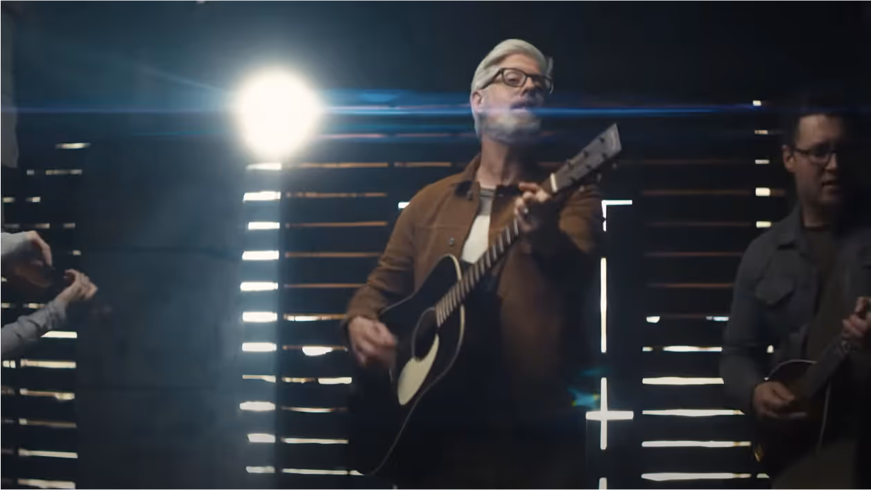 An image of a man with white hair performing a song with an acoustic guitar.