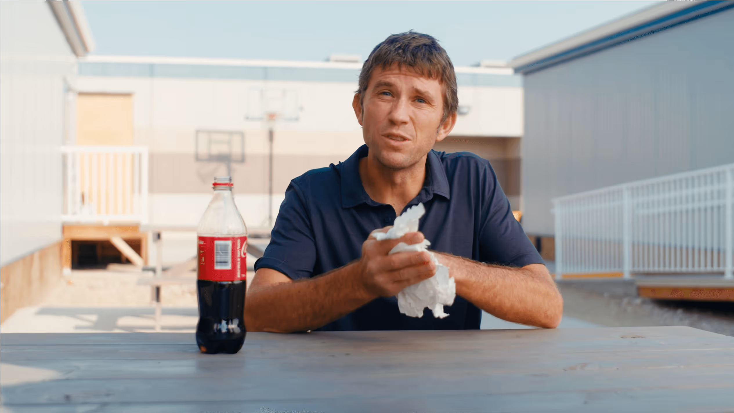 An image of a middle aged man sitting at a picnic table with a bottle of coke.