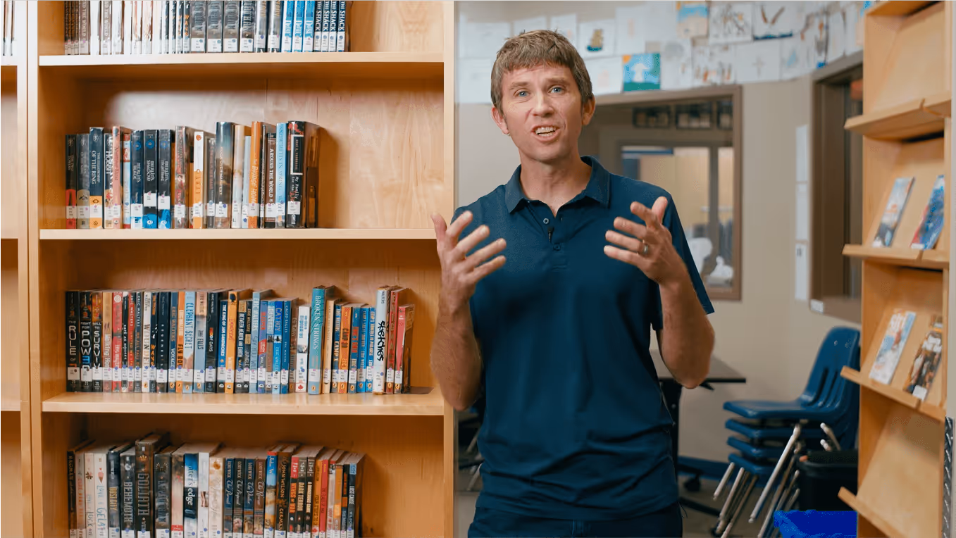 An image of a man leaning against a bookshelf expressively talking to the camera.