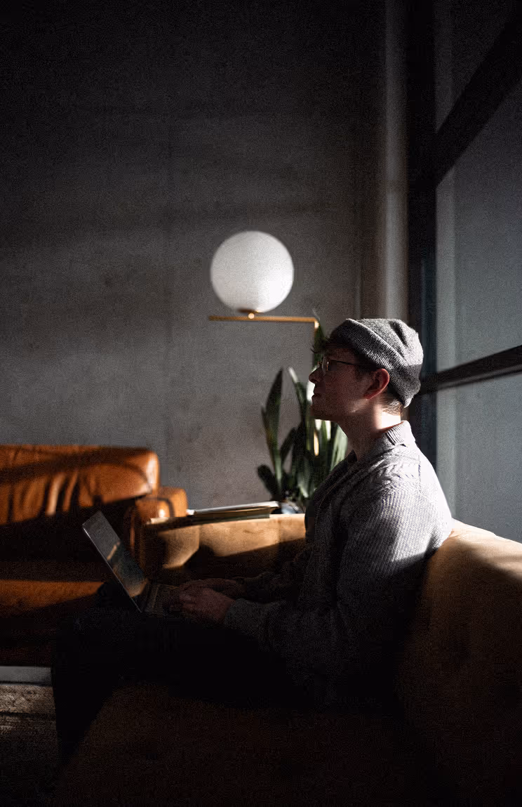A young man sitting on a velour couch with light shining on his back. 