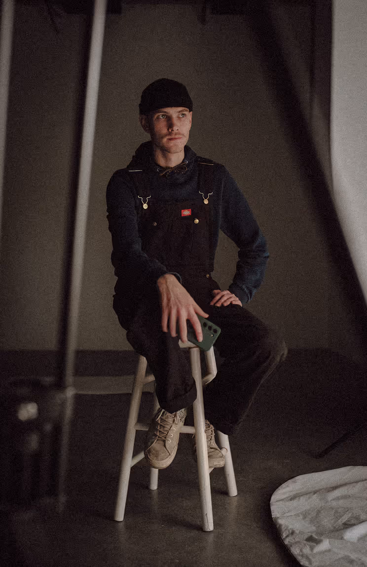 A young man in overalls sitting on a stool in a film studio.