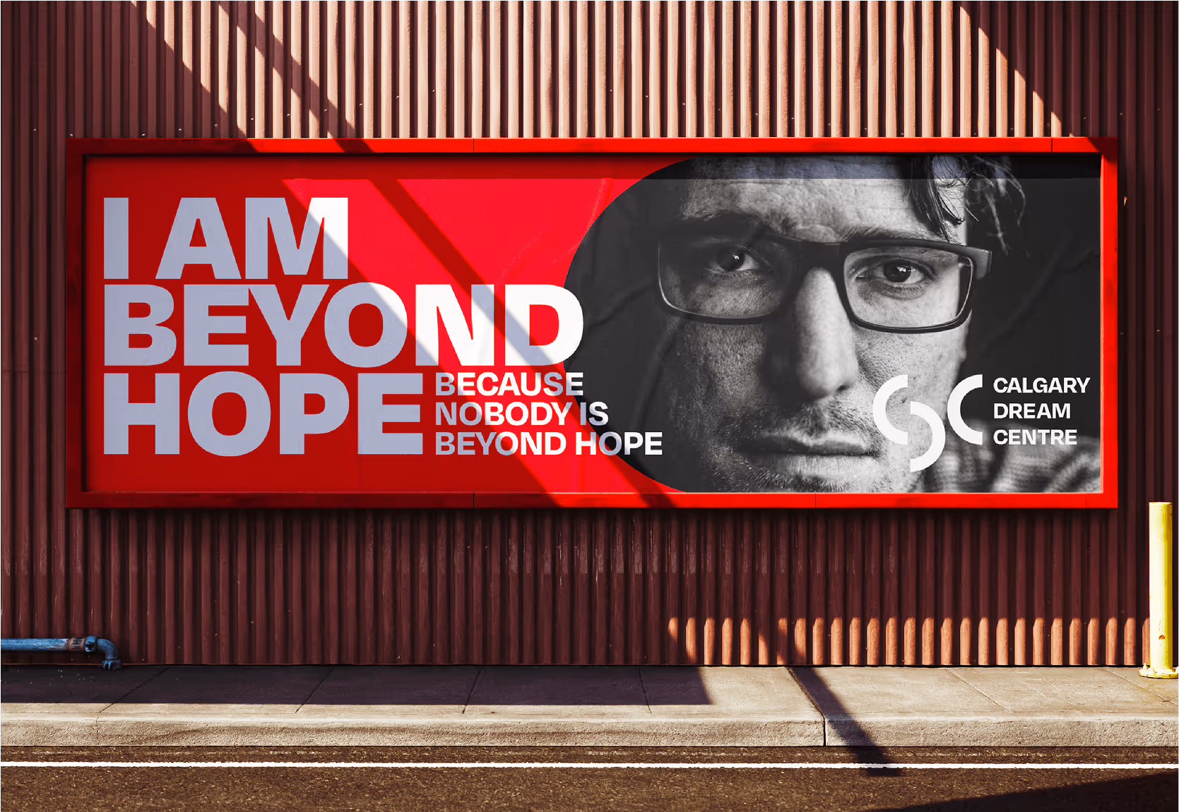 Billboard with red background featuring a close-up black and white photo of a man wearing glasses and white text saying 'I AM BEYOND HOPE BECAUSE NOBODY IS BEYOND HOPE' and the Calgary Dream Centre logo.