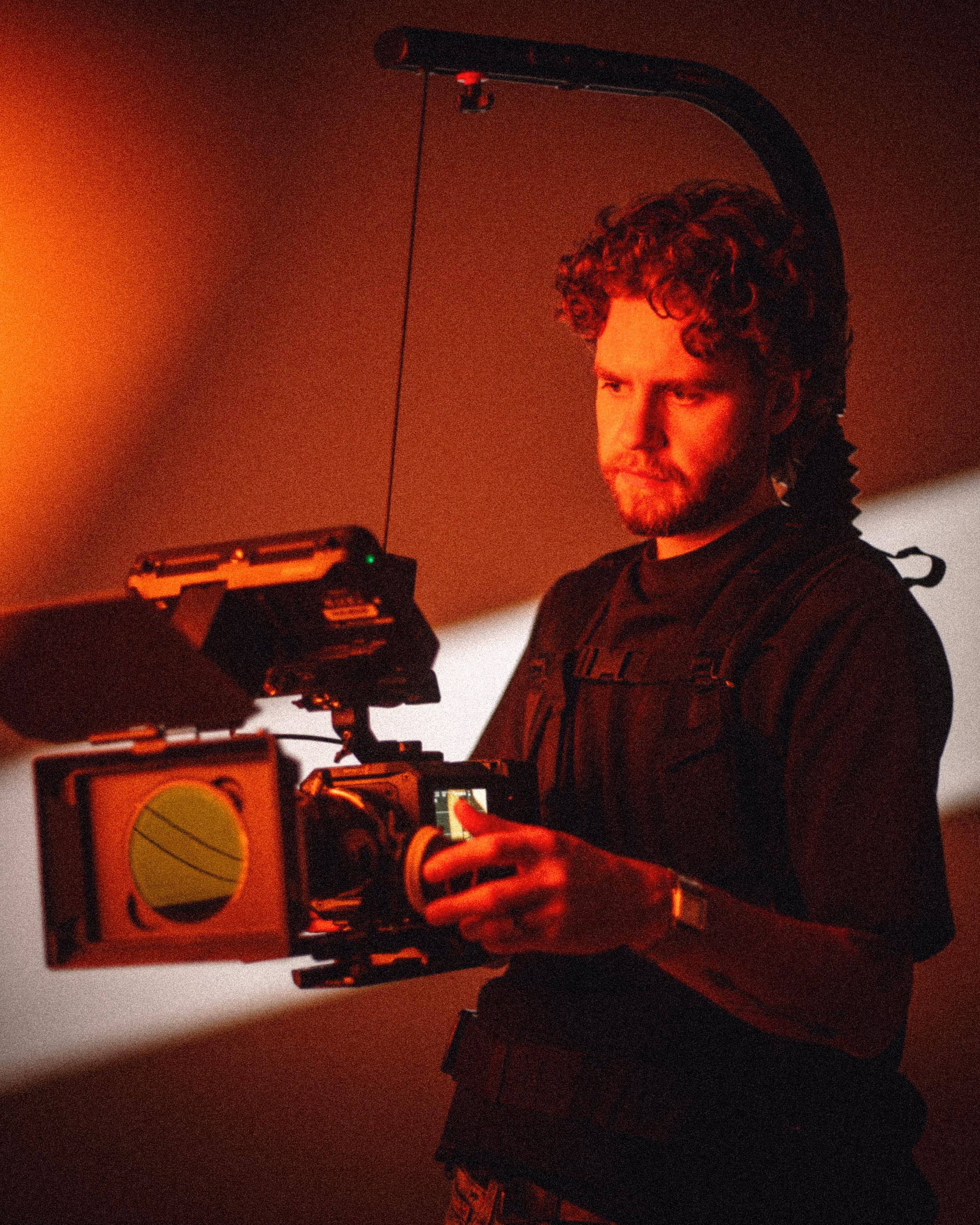 Man with curly hair operating a professional video camera rig with a matte box in warm red lighting.