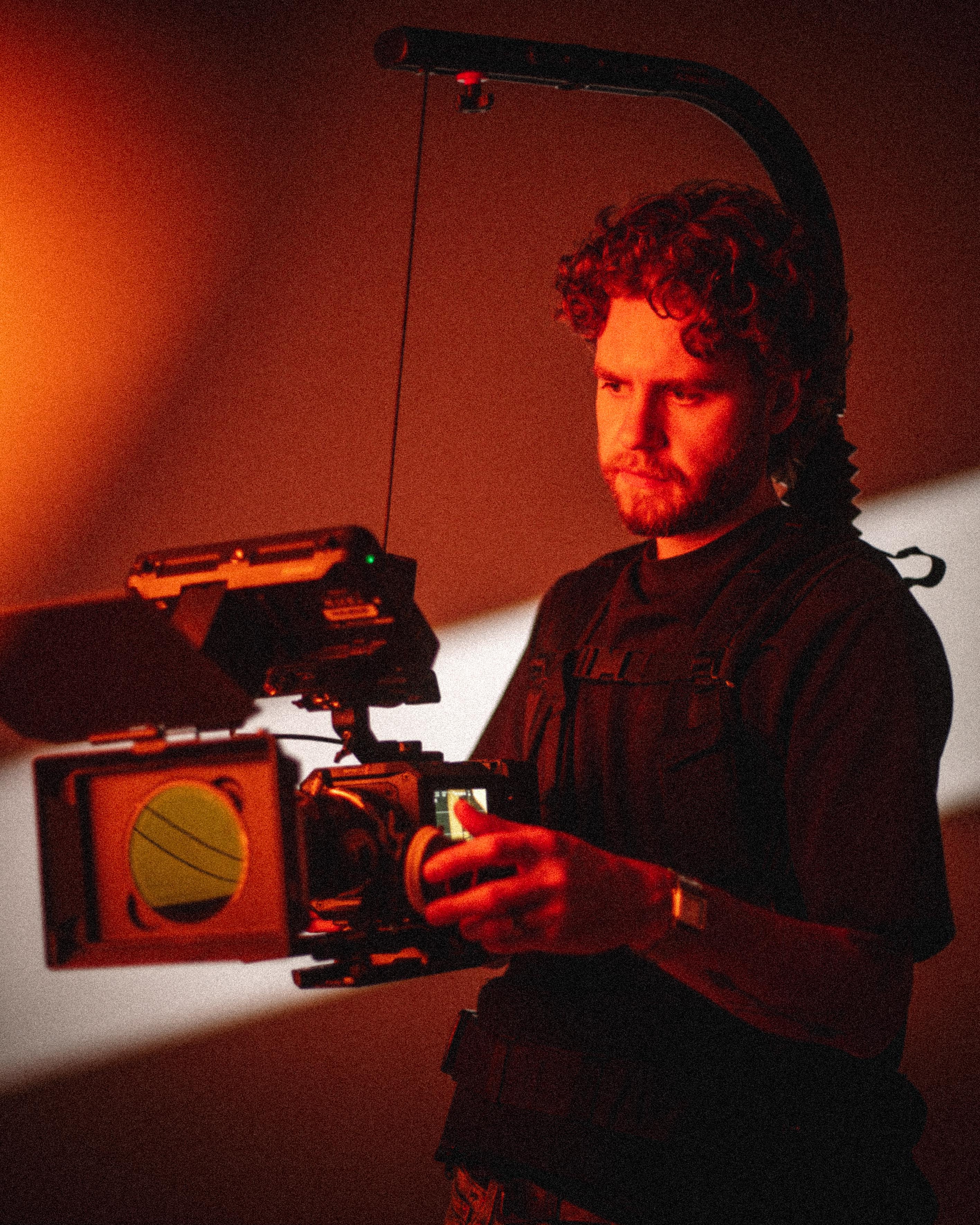 Man with curly hair operating a professional video camera rig with a matte box in warm red lighting.