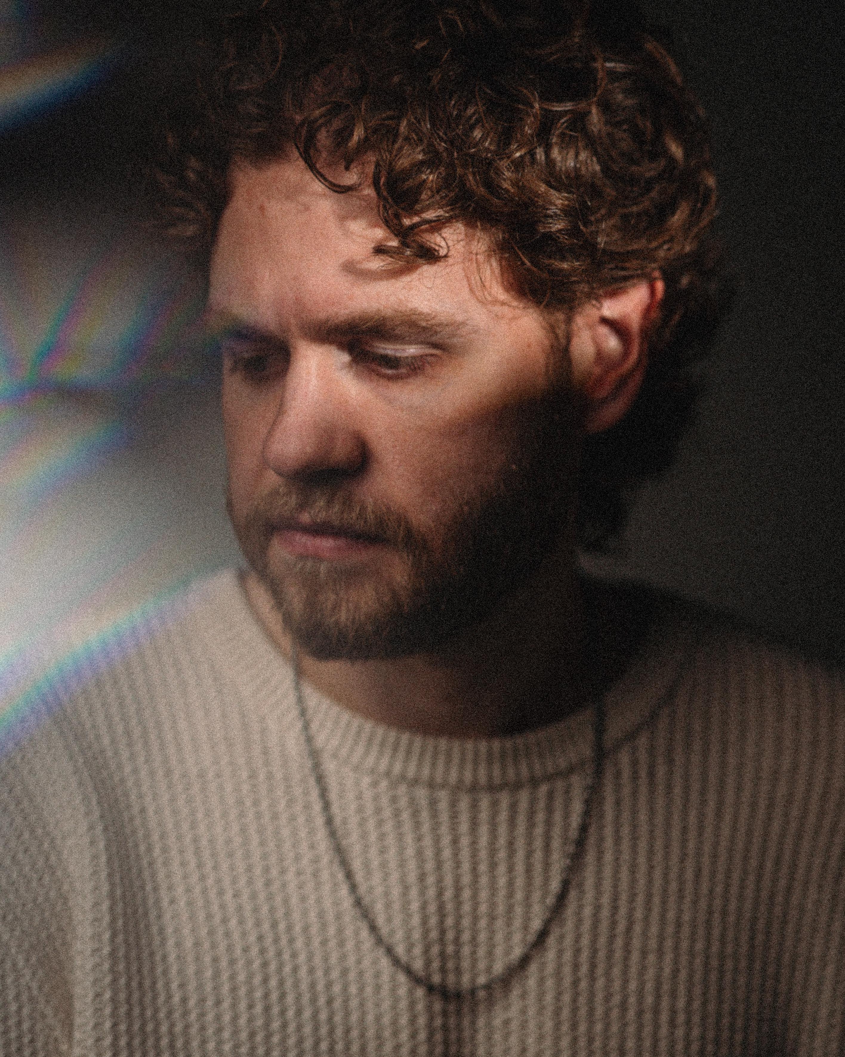 Portrait of a man with curly hair and beard, wearing a beige knit sweater and a chain necklace, looking downward.