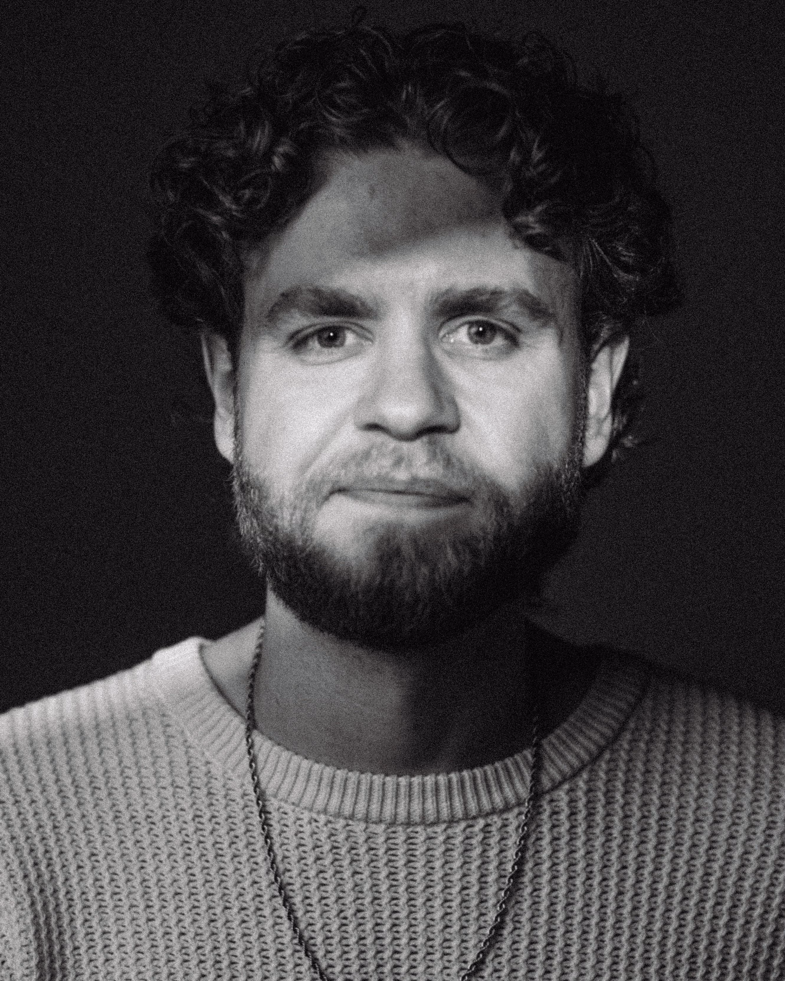 Black and white close-up portrait of a man with curly hair and a beard wearing a knit sweater and chain necklace.