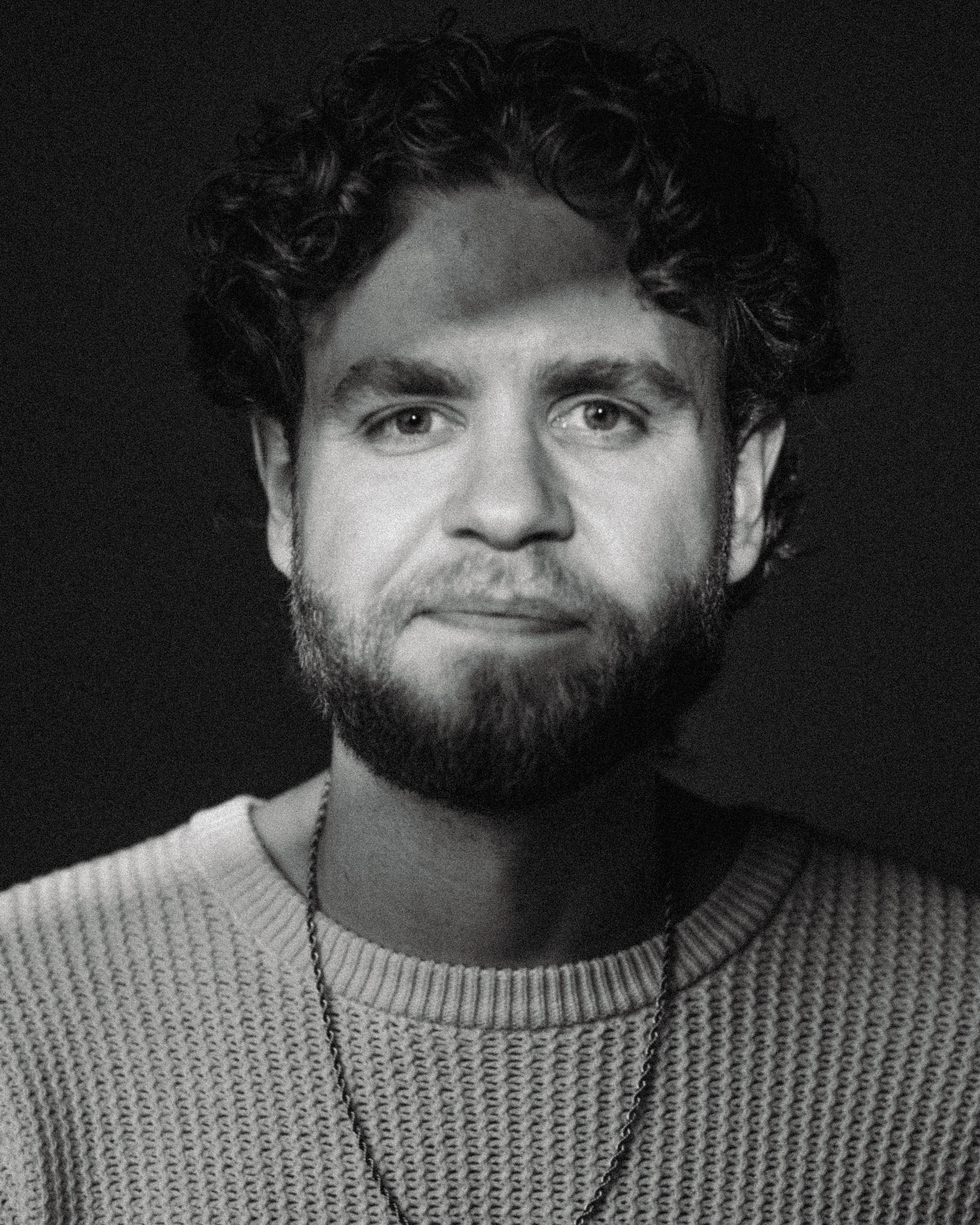 Black and white close-up portrait of a man with curly hair and a beard wearing a knit sweater and chain necklace.