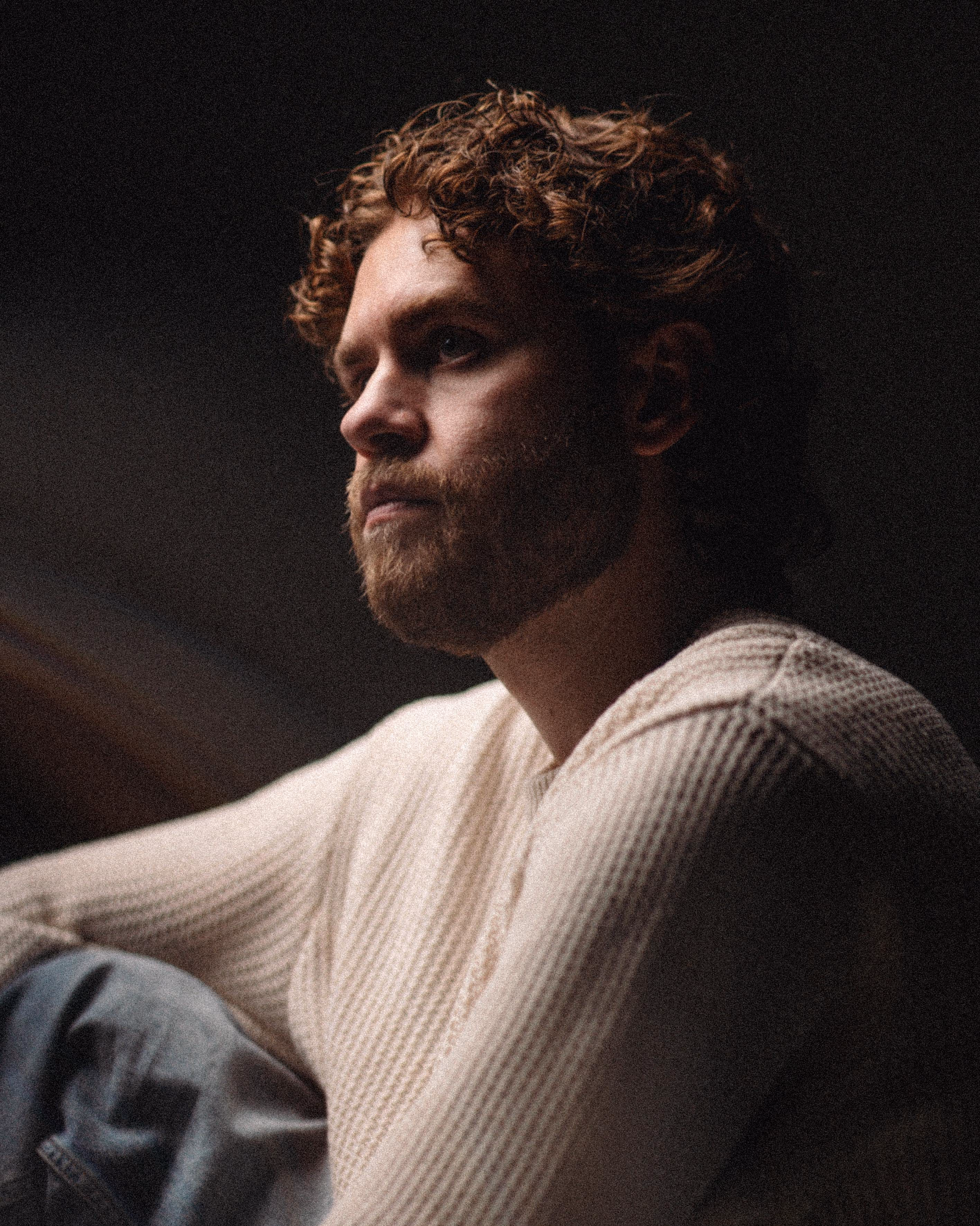 Thoughtful man with curly hair and beard wearing a cream-colored knit sweater looking to the side in low light.