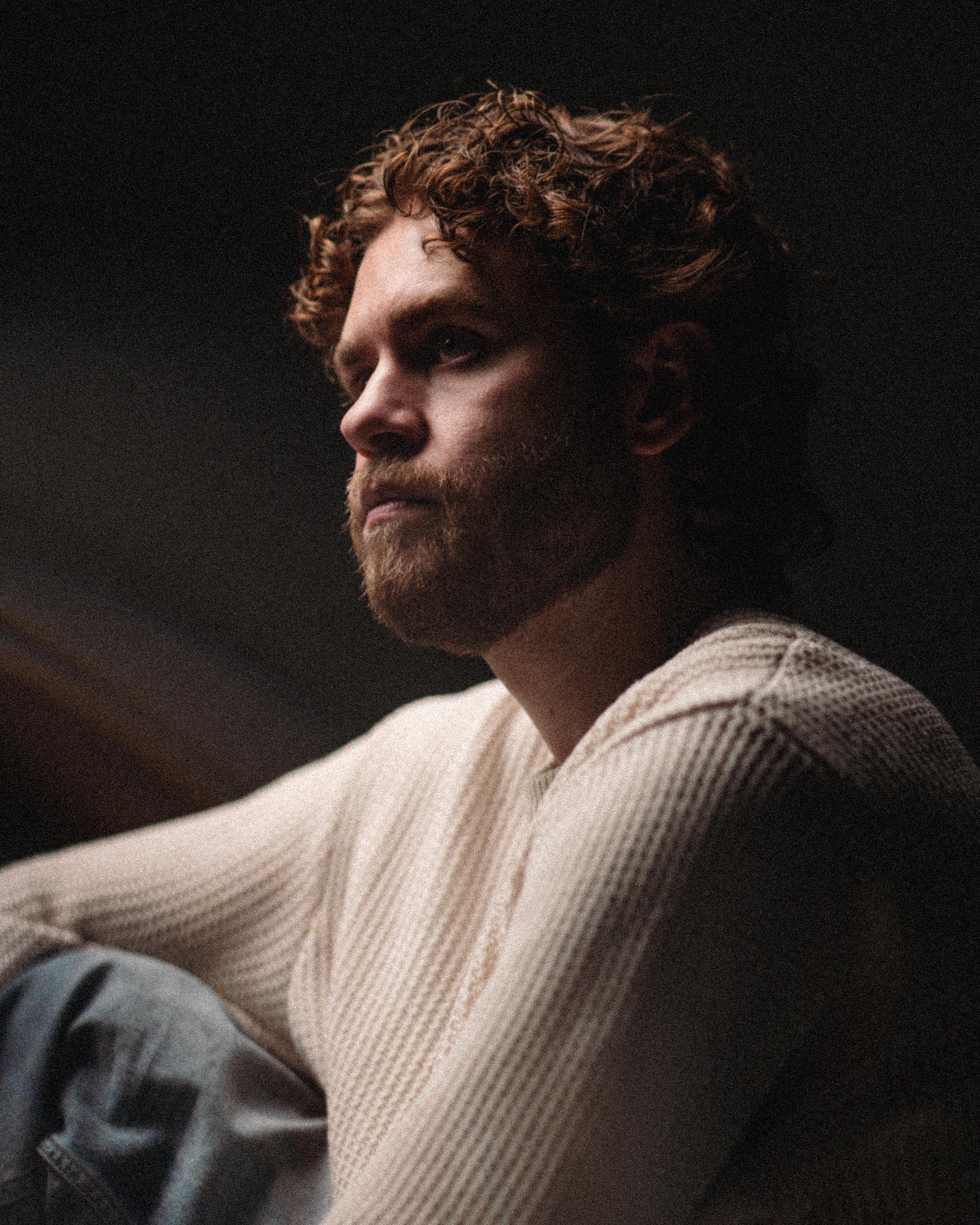 Thoughtful man with curly hair and beard wearing a cream-colored knit sweater looking to the side in low light.