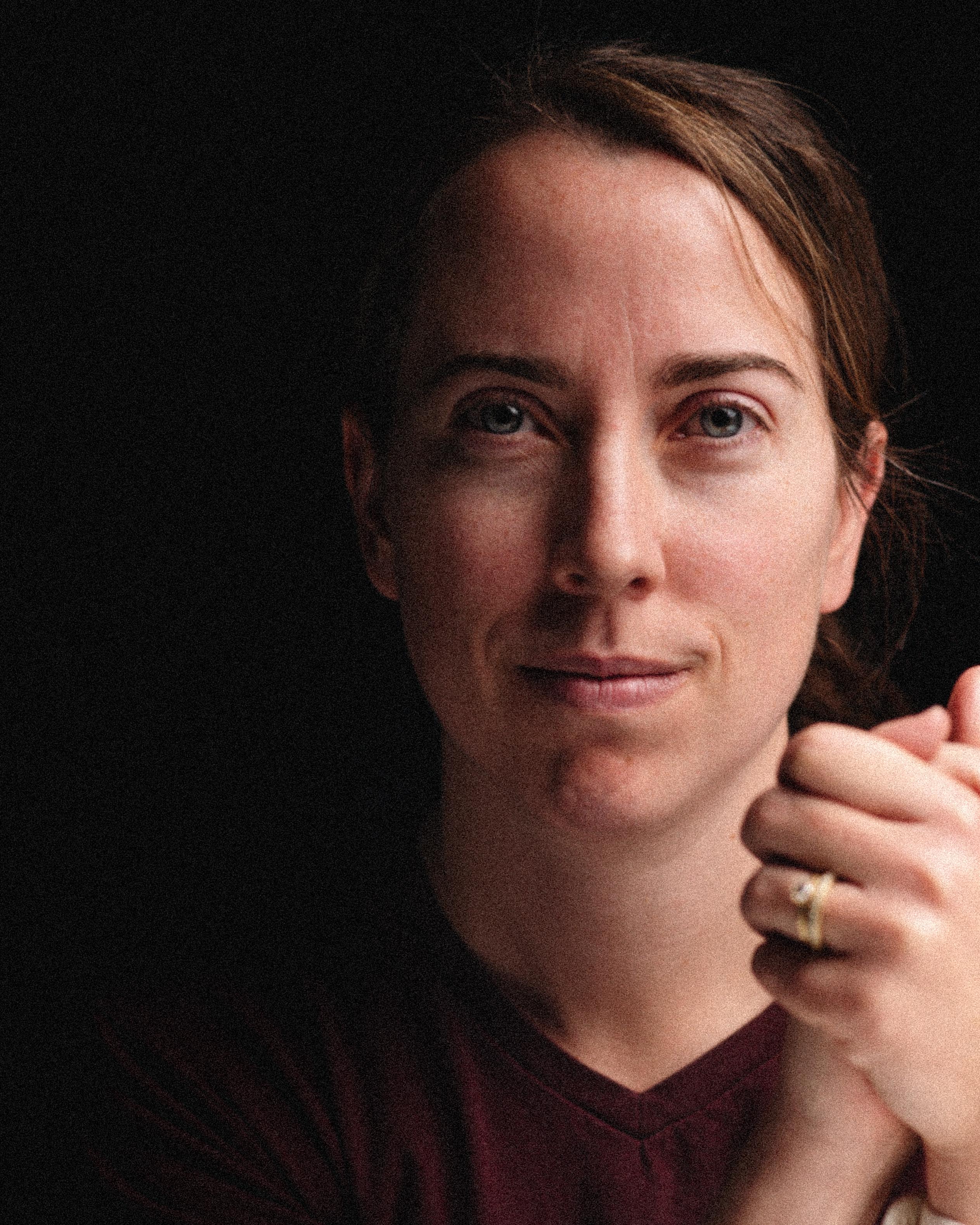 Close-up portrait of a woman with light brown hair and a ring on her hand, against a dark background.