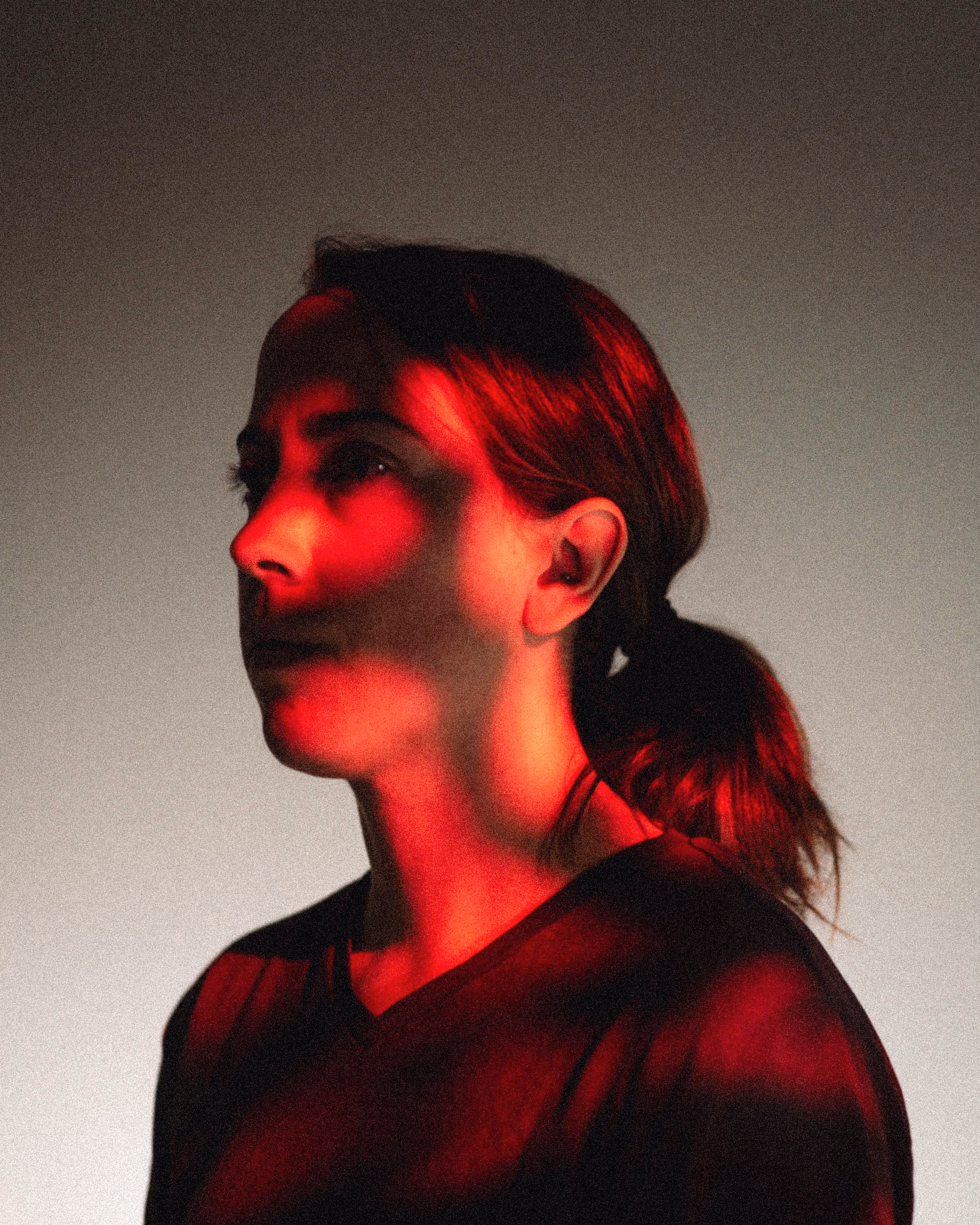 Portrait of a woman with her hair tied back, illuminated by dramatic red lighting against a neutral background.