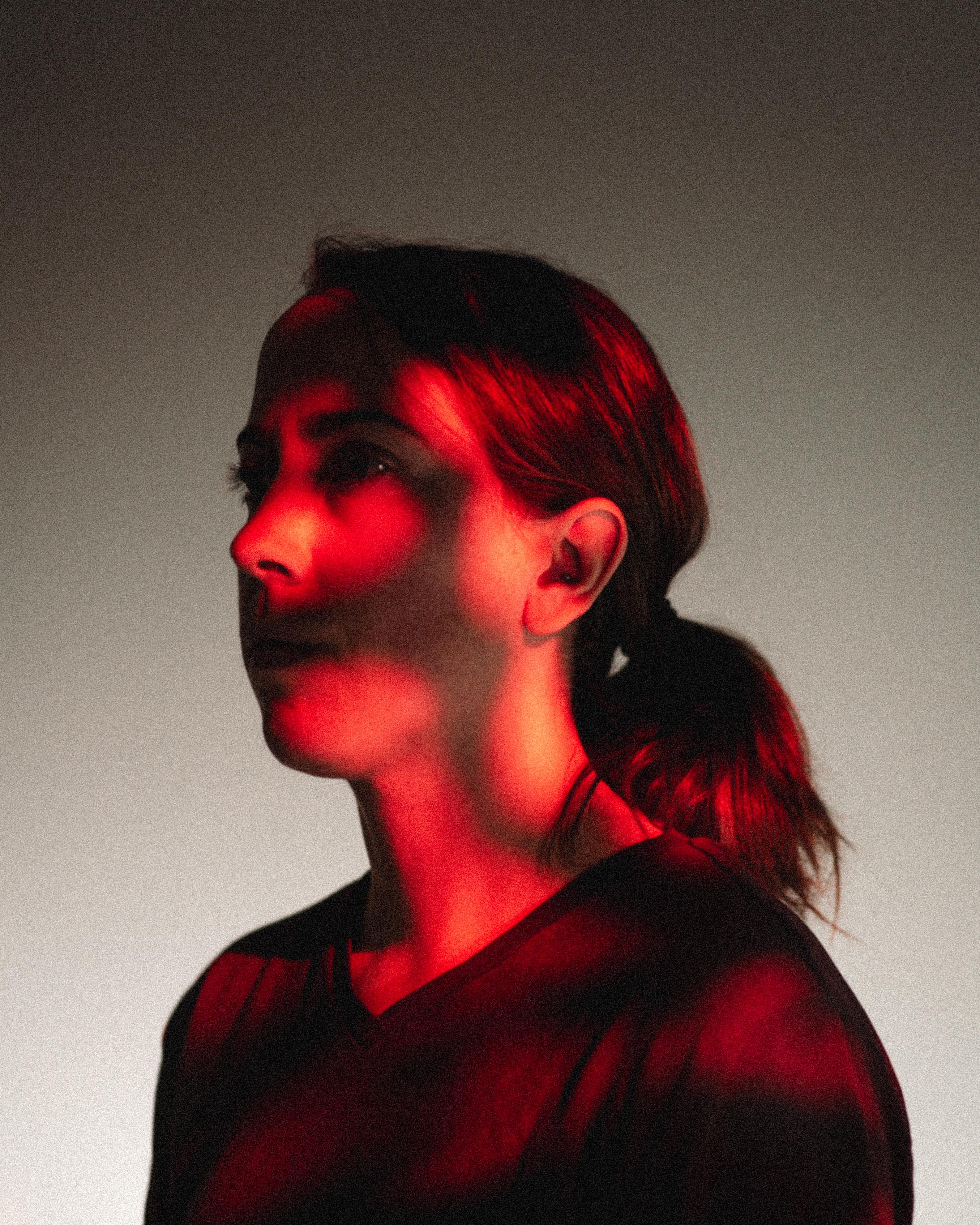 Portrait of a woman with her hair tied back, illuminated by dramatic red lighting against a neutral background.
