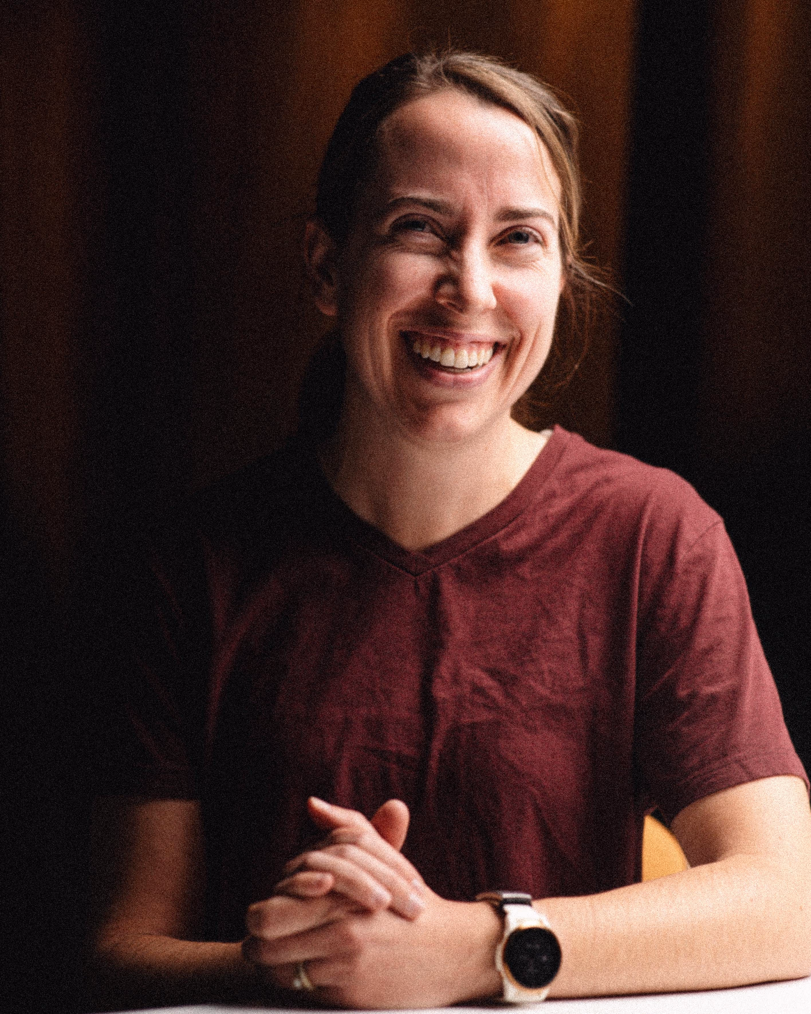 Smiling woman with light skin and brown hair tied back, wearing a maroon shirt and a smartwatch, sitting at a table with hands clasped.