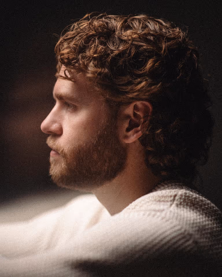 Profile portrait of a man with curly hair and beard wearing a white textured sweater against a dark background.