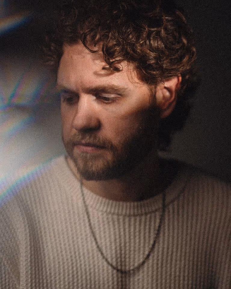 Man with curly hair and beard wearing a light knitted sweater and a chain necklace looking down thoughtfully.
