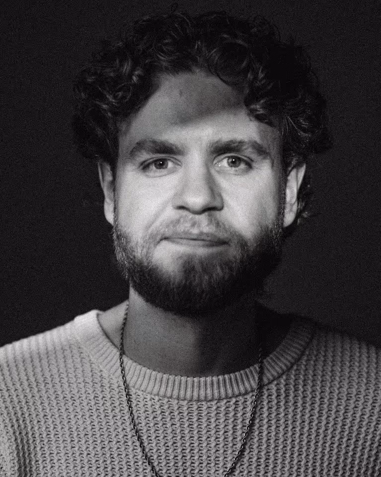 Black and white portrait of a young man with curly hair, a beard, and a knit sweater, looking directly at the camera.