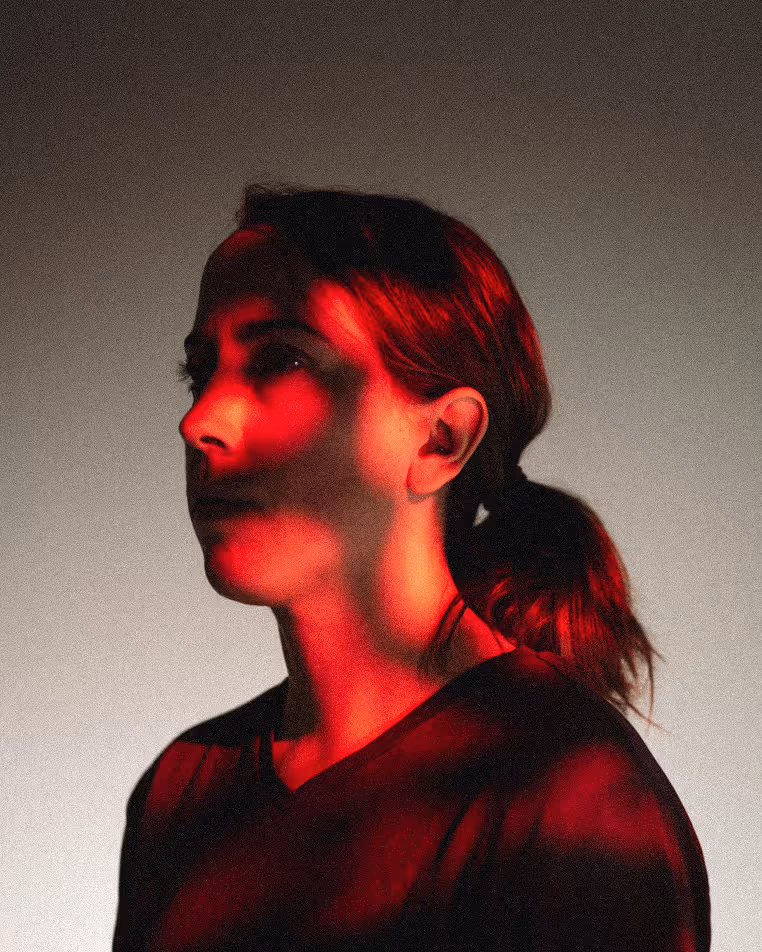 Portrait of a woman with her hair tied back, illuminated by dramatic red and black lighting against a neutral background.