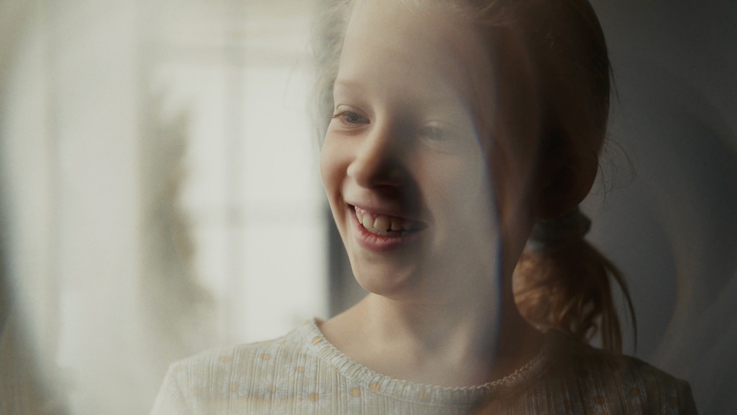 Smiling young girl with a ponytail looking out softly lit window.