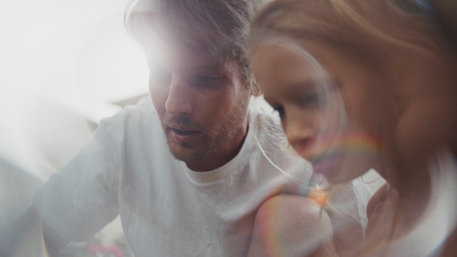 Man with light brown hair and beard wearing a white t-shirt leaning in close to his young daughter in a softly lit setting.