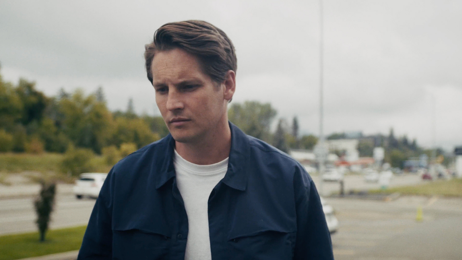 Young man with brown hair wearing a navy jacket and white shirt walking with a pensive expression.