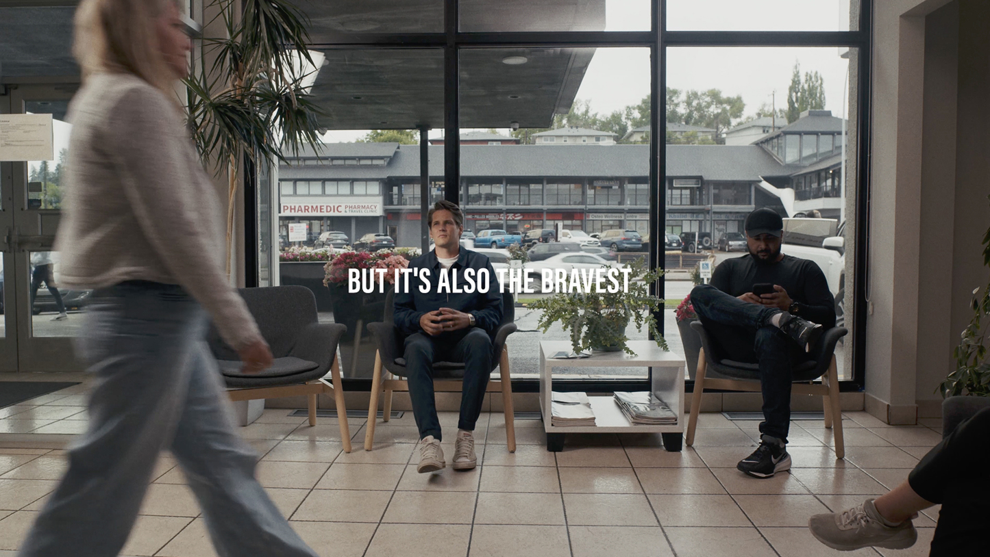 Two men sit in chairs inside the CDC waiting area by large windows, with a woman walking past in the foreground and the text 'BUT IT'S ALSO THE BRAVEST' overlaid.