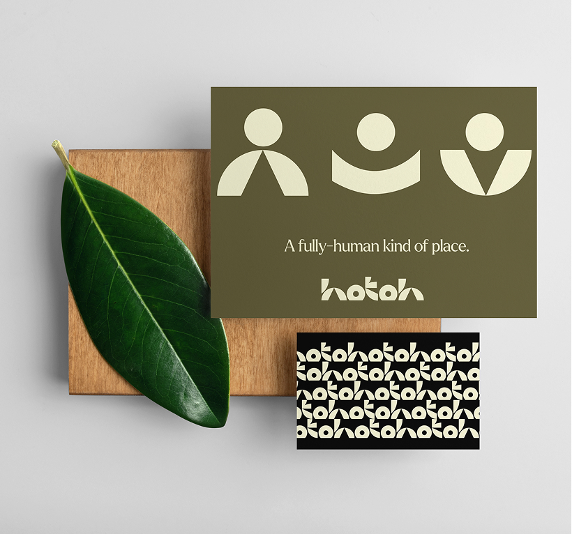 Two cards on wooden board next to a green leaf; the larger card features three abstract human shapes and text 'A fully-human kind of place.' with 'hotoh' logo, smaller card has repeating 'hotoh' logo pattern on black background.