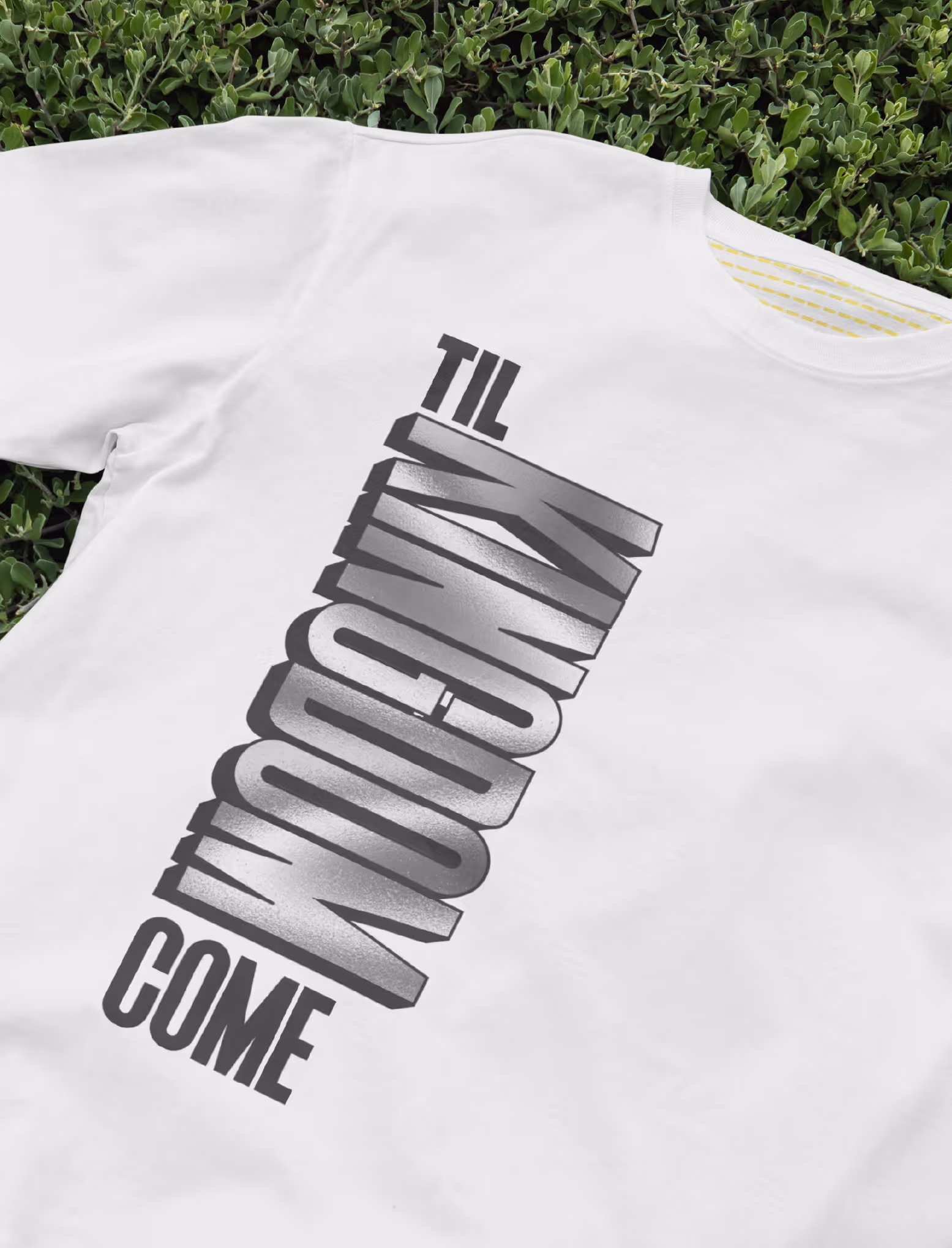 White t-shirt with large diagonal text reading 'TIL TOMORROW COME' laid on green foliage.