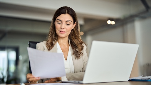 woman sitting at a desk looking over a document and a laptop