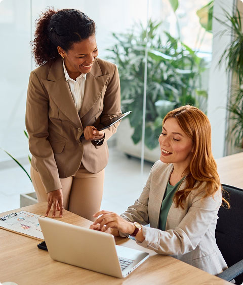 woman standing and holding a tablet, while talking to a seated woman working at a laptop