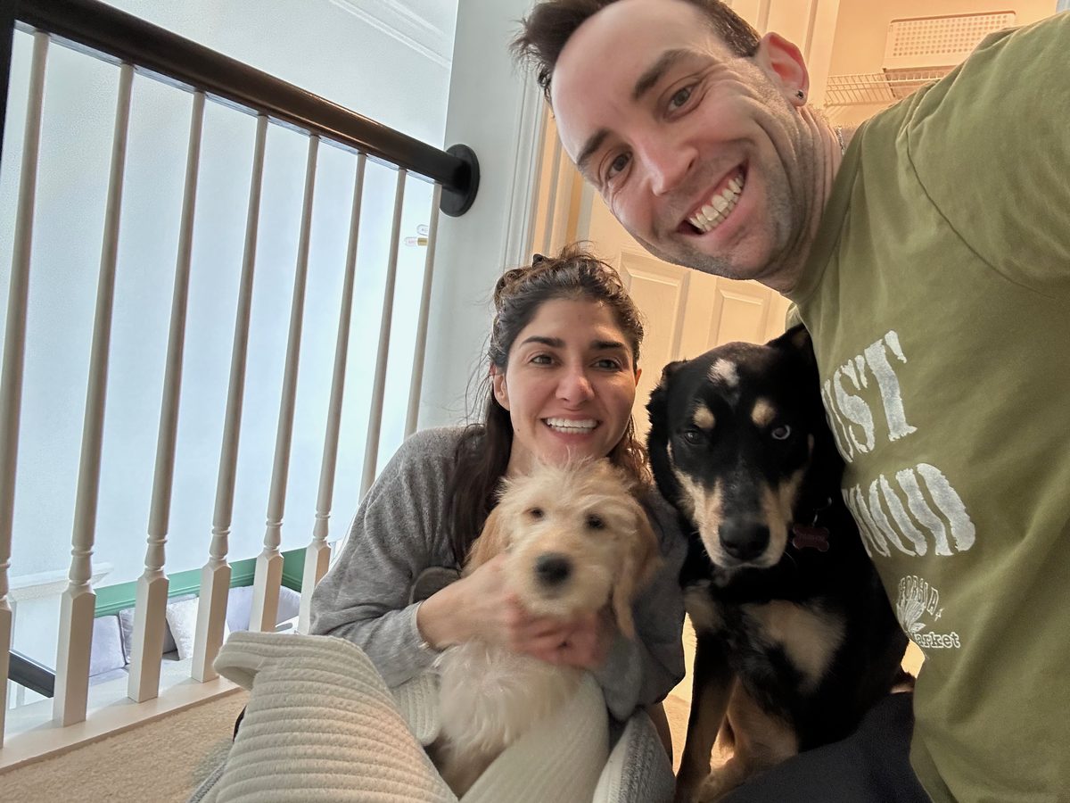 Roxanna and her husband Ian indoors sitting on carpet with two dogs, one black and tan and one light-colored fluffy dog.