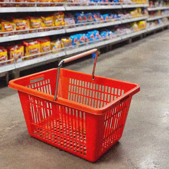 Roter Einkaufskorb mit Metallgriffen auf dem Boden eines Supermarkts vor Regalen mit verschiedenen Produkten.