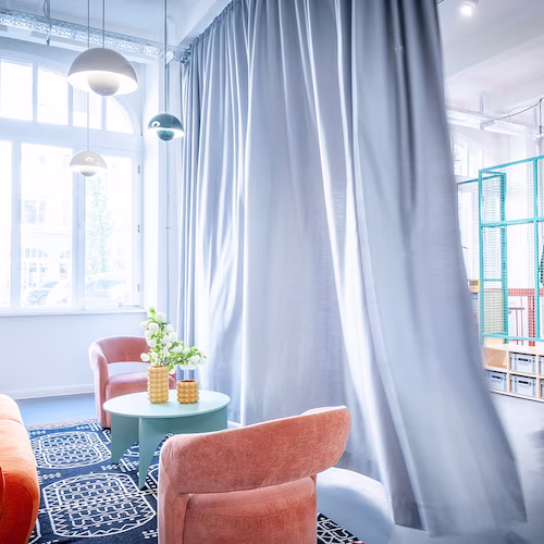 Helles Wohnzimmer mit rosa Sesseln, blauem Teppich, rundem Tisch und großen grauen Vorhängen.