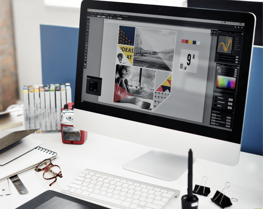 Desk setup with an iMac displaying a graphic design project, surrounded by art markers, glasses, and office supplies.