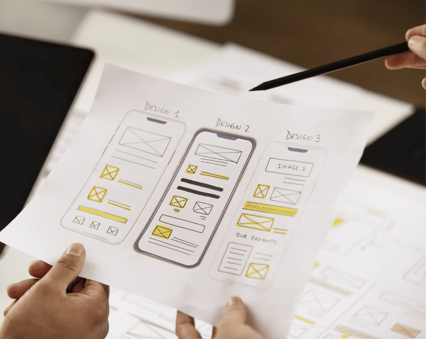 Hands holding a printed paper with three hand-drawn mobile app design wireframes labeled Design 1, Design 2, and Design 3, with a pen pointing at Design 2.