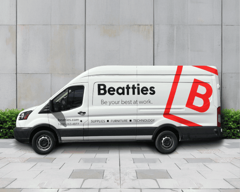 White delivery van with black lower trim parked on stone pavement in front of a concrete wall with green shrubbery, featuring large red 'B' logo and black text 'Beatties Be your best at work. supplies furniture technology' on the side.