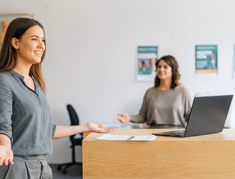 Deux femmes souriantes discutant dans un bureau avec un ordinateur portable posé sur une table en bois.