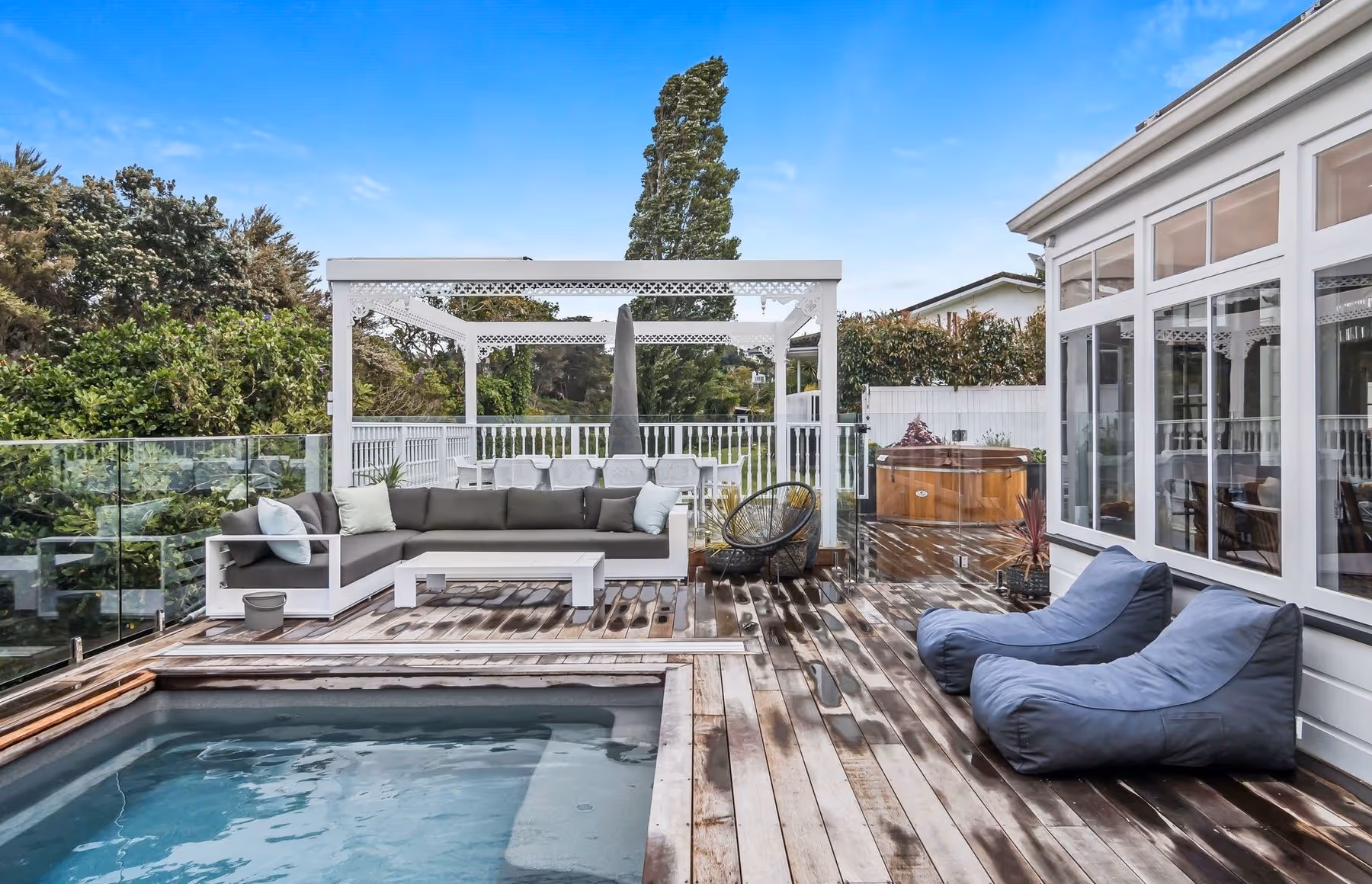 A stylish outdoor deck with a hot tub, pool, seating area, and lush greenery in the backdrop under a clear blue sky.