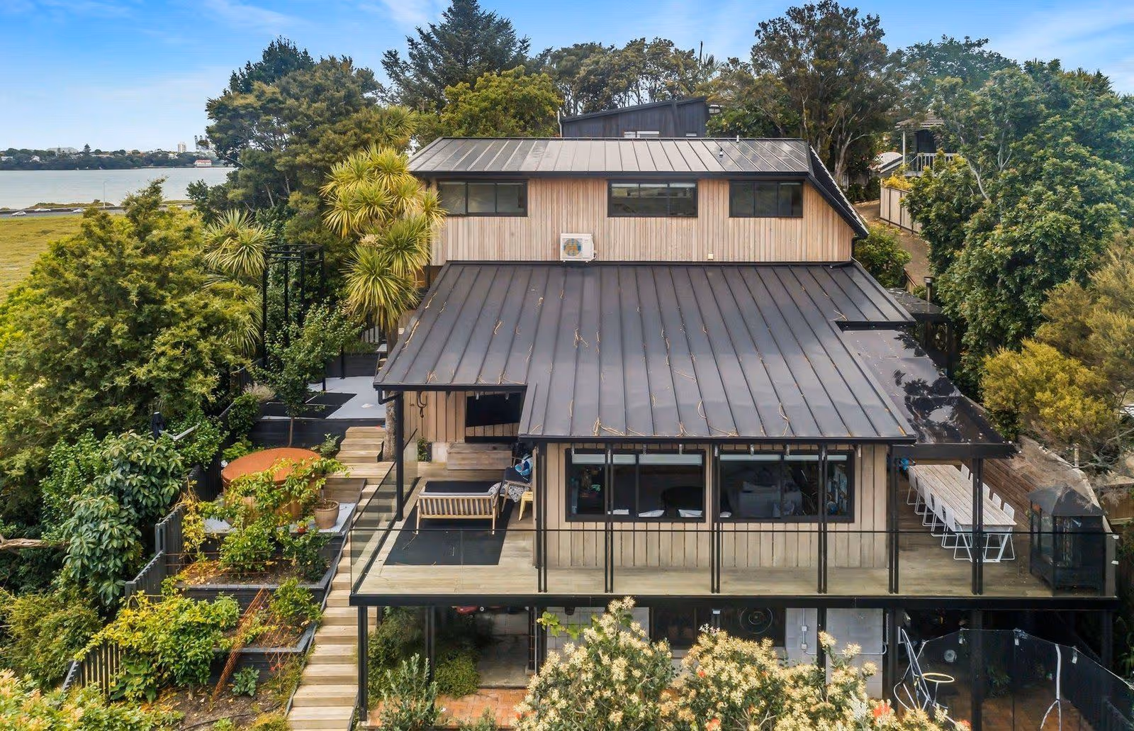 A modern, wooden house with a black metal roof, surrounded by lush greenery and overlooking a serene water view.
