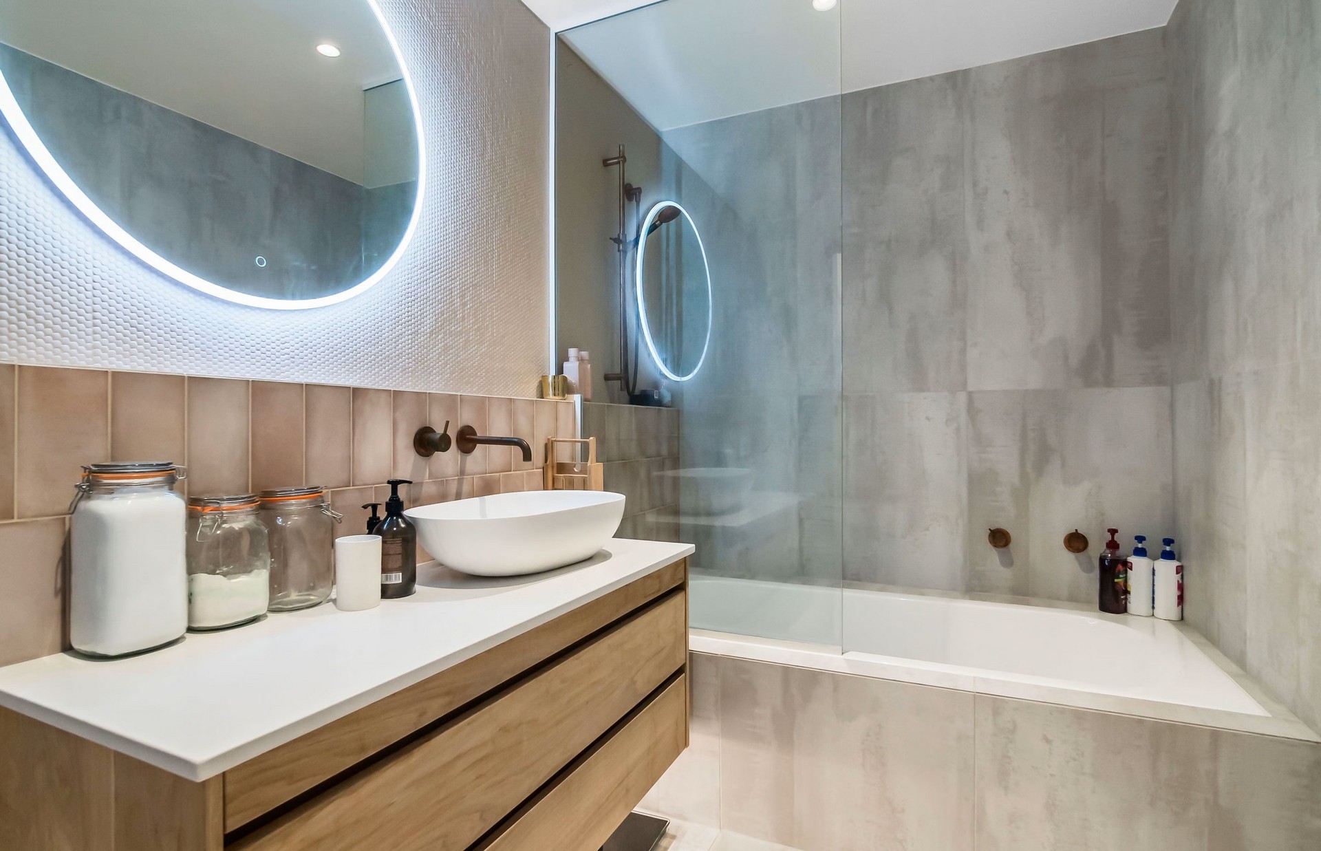 Modern bathroom featuring a circular illuminated mirror, a sleek basin, wooden cabinetry, and a glass shower beside a tub.