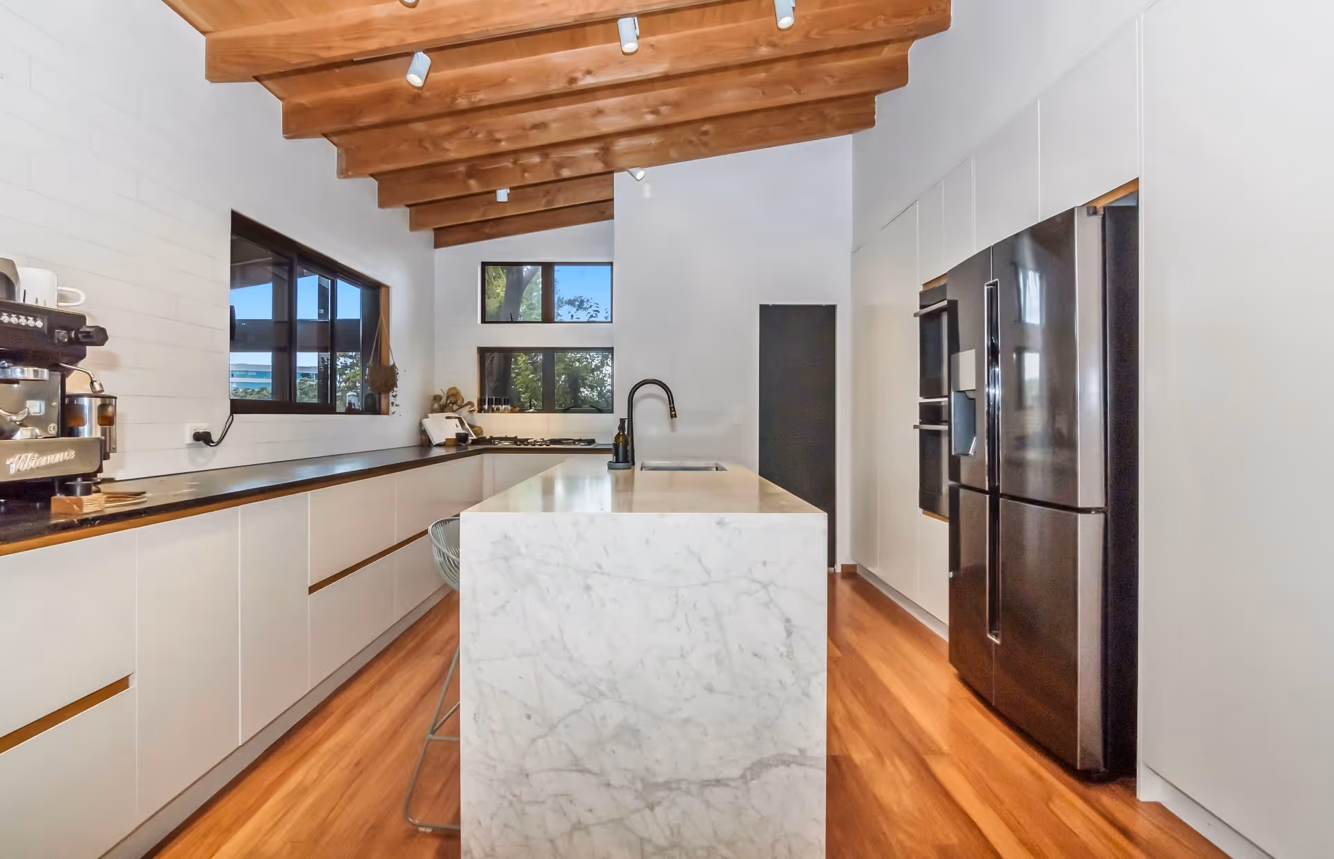Modern kitchen featuring a marble island, wooden beams, sleek cabinetry, and large windows allowing natural light.